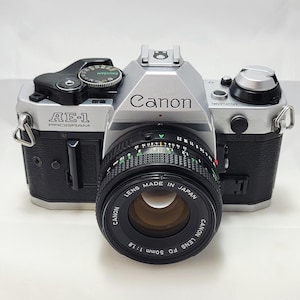 May include: A silver and black Canon AE-1 Program camera with a 50mm lens. The camera body has the Canon logo and AE-1 Program text. The lens is black with white lettering, marked "LENS MADE IN JAPAN".