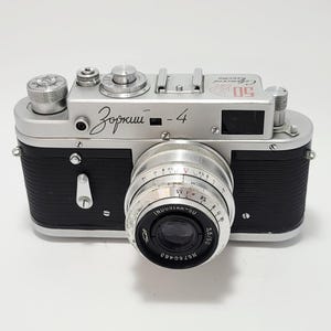 May include: A vintage silver and black Zorki-4 camera with a lens. The camera has several dials and a viewfinder. The lens is marked with the text "INDUSTAR-50 3.5/50". The camera is on a white background.