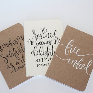 May include: Three small notebooks with inspirational quotes written in calligraphy. The notebooks are brown with white and black lettering. The quotes are "He makes my feet like deer's feet", "He rescued me because He delights in me", and "Free indeed".