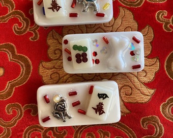 Pode incluir: Três peças decorativas retangulares brancas com peças de Mahjong e amuletos. Cada peça tem um amuleto diferente: um cão, uma rosa e um osso. As peças são decoradas com contas vermelhas, azuis e amarelas e confetes iridescentes.