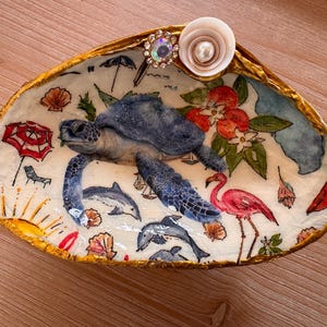 May include: A decorative clam shell dish with a gold-painted rim, featuring a central sea turtle illustration surrounded by dolphins, a pink flamingo, a sun, and beach umbrellas. A small white flower with a pearl and rhinestone center is attached.
