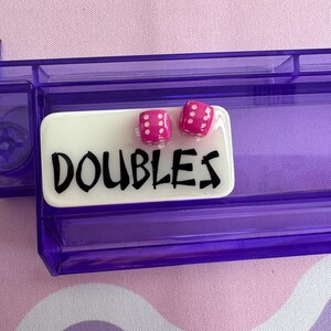 May include: A purple dice tray with a white rectangular label that reads "DOUBLES" in black lettering. Two pink dice with white dots are on top of the label. The tray is made of clear plastic.