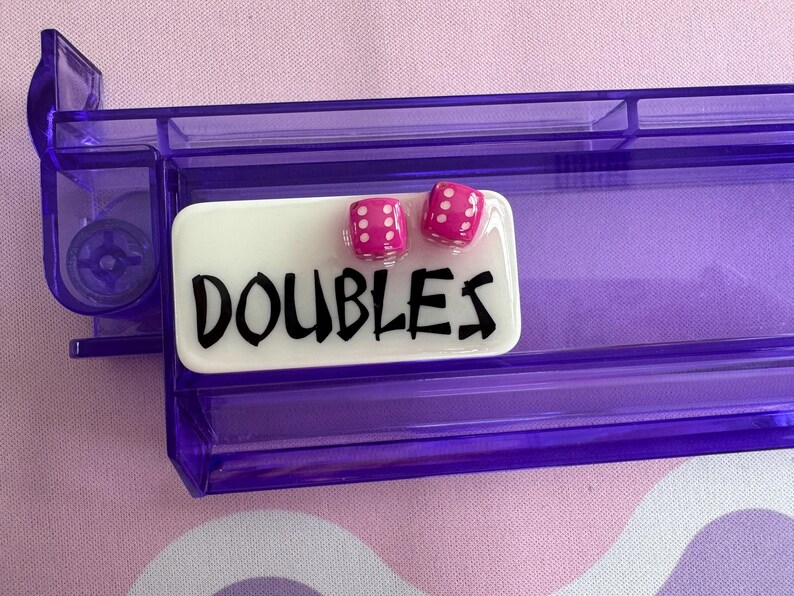 May include: A purple dice tray with a white rectangular label that reads "DOUBLES" in black lettering. Two pink dice with white dots are on top of the label. The tray is made of clear plastic.