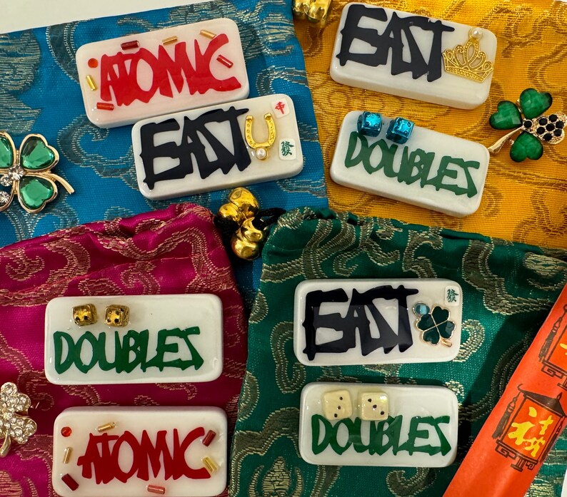 May include: A collection of white rectangular resin pieces with various designs. Some feature the words "ATOMIC", "EAST", and "DOUBLES" in bold, graffiti-style lettering. Embellishments include dice, a horseshoe, and a crown.