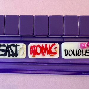 May include: A purple Mahjong tile rack with three white tiles. The tiles have the words "EAT", "ATOMIC", and "DOUBLES" written on them. Two pink dice are on the "DOUBLES" tile. The background is a light pink.