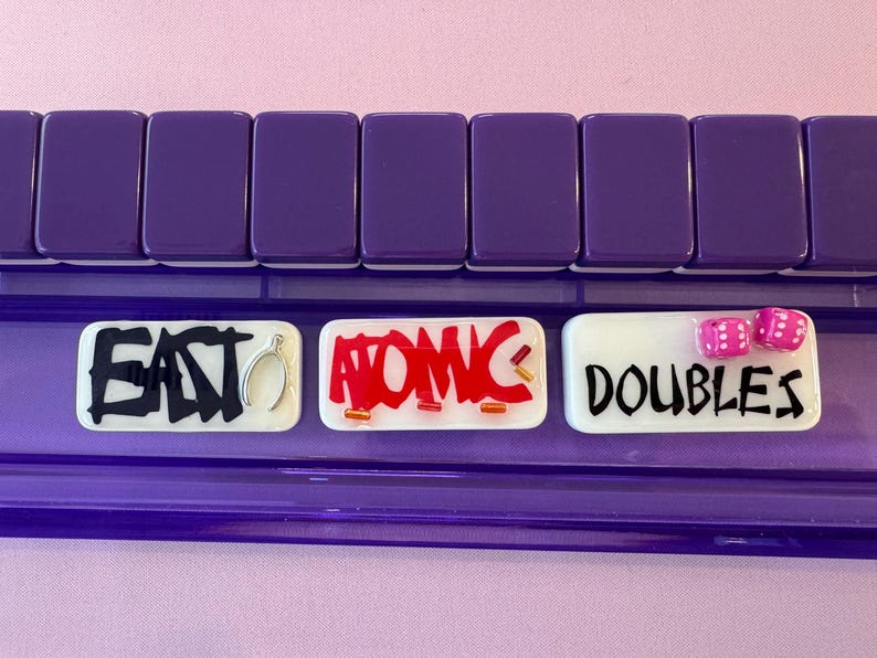 May include: A purple Mahjong tile rack with three white tiles. The tiles have the words "EAT", "ATOMIC", and "DOUBLES" written on them. Two pink dice are on the "DOUBLES" tile. The background is a light pink.