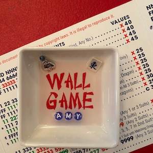 May include: A white square dish with the words "WALL GAME" in red and the name "AMY" in blue beads. The dish contains a small charm and a tile with a Chinese character. The dish sits on a red surface with game instructions.