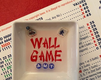Puede incluir: Un plato cuadrado blanco con las palabras "WALL GAME" en rojo y el nombre "AMY" en cuentas azules. El plato contiene un pequeño amuleto y una ficha con un carácter chino. El plato está sobre una superficie roja con instrucciones del juego.