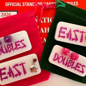 May include: Two red velvet pouches with white rectangular game pieces. One pouch has pieces that say "DOUBLES" and "EAST" in purple, with purple dice and a money bag charm. The other pouch has pieces that say "DOUBLES" and "EAST" in purple, with pink dice and a horseshoe charm.