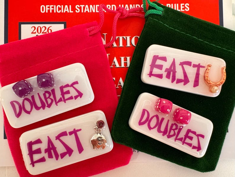 May include: Two red velvet pouches with white rectangular game pieces. One pouch has pieces that say "DOUBLES" and "EAST" in purple, with purple dice and a money bag charm. The other pouch has pieces that say "DOUBLES" and "EAST" in purple, with pink dice and a horseshoe charm.