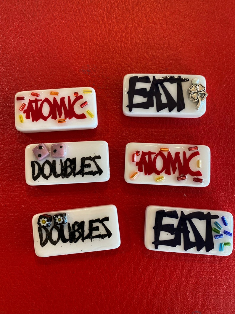 May include: Six white rectangular glass pieces with black and red text and decorative elements. The words "ATOMIC", "DOUBLES", and "EAT" are displayed. Some pieces have dice or a four-leaf clover.