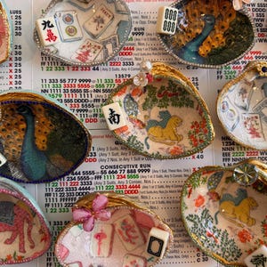 May include: A collection of decorative seashell dishes, each uniquely adorned with painted designs and embellished with Mahjong tiles. The shells feature images of peacocks, floral patterns, and other motifs, with gold-painted edges.