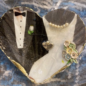 May include: Heart-shaped wedding decoration featuring a tuxedo and a wedding dress design. The decoration has a gold trim and includes a small bouquet and a charm that says "Ring Bearer".