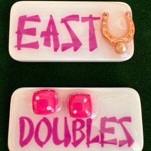 May include: Two white rectangular game pieces with pink text and accents. One piece reads "EAST" with a gold horseshoe and pearl. The other reads "DOUBLES" with two pink dice. The dice have white dots.