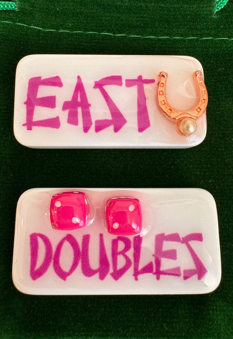 May include: Two white rectangular game pieces with pink text and accents. One piece reads "EAST" with a gold horseshoe and pearl. The other reads "DOUBLES" with two pink dice. The dice have white dots.