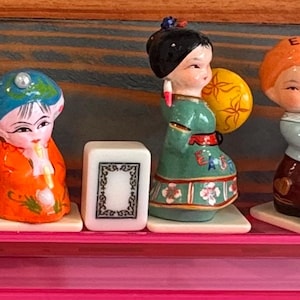 Può includere: Una collezione di dieci statuette in ceramica disposte in fila su uno scaffale rosa. Le figure presentano vari abiti colorati, alcune tengono ventagli o accessori, con diverse parti che mostrano la parola EAST stampata sui vestiti.