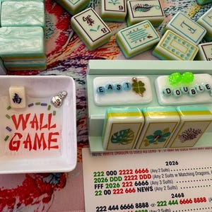 May include: A Mahjong game set with various tiles, dice, and game accessories. The set includes tiles with Chinese characters and symbols, along with the words "WALL GAME" in red. The set is arranged on a colorful floral patterned surface.