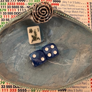 May include: A blue-swirled dish contains a silver spiral ring, a white Mahjong tile with black characters, and two blue dice with white dots. The dish is edged with small, clear beads. A printed list of numbers is in the background.