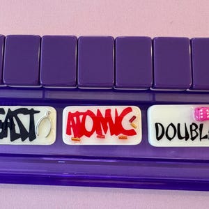 May include: A purple game board with three white rectangular tiles. The tiles have the words "EAST", "ATOMIC", and "DOUBLES" written on them in black and red. Two pink dice are on the "DOUBLES" tile.