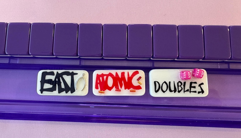 May include: A purple game board with three white rectangular tiles. The tiles have the words "EAST", "ATOMIC", and "DOUBLES" written on them in black and red. Two pink dice are on the "DOUBLES" tile.