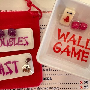 May include: A red velvet pouch and a white tray with the text "WALL GAME" in red. The tray contains a Mahjong tile, two dice, and a gold horseshoe. Two white trays with the text "DOUBLES" and "EAST" in purple are also present.
