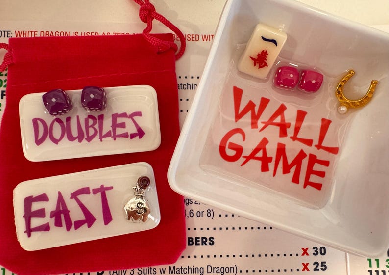 May include: A red velvet pouch and a white tray with the text "WALL GAME" in red. The tray contains a Mahjong tile, two dice, and a gold horseshoe. Two white trays with the text "DOUBLES" and "EAST" in purple are also present.