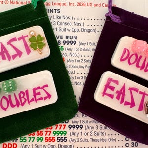 May include: Two small velvet pouches, one green and one purple, each holding game pieces. The green pouch has a white tile with "EAST" in pink and green dice. The purple pouch has a tile with "DOUBLES" and pink dice.