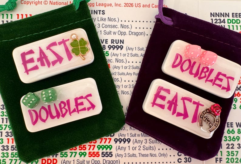May include: Two small velvet pouches, one green and one purple, each holding game pieces. The green pouch has a white tile with "EAST" in pink and green dice. The purple pouch has a tile with "DOUBLES" and pink dice.