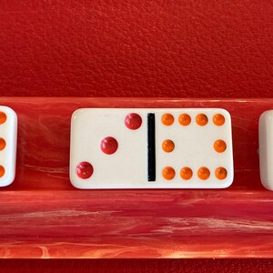 May include: Three white dominoes with black center lines and colored dots. The first domino has orange dots, the second has red dots, and the third has blue and green dots. The dominoes are displayed on a red and pink surface.
