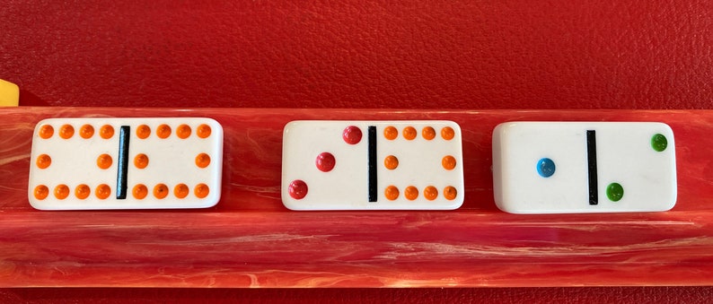 May include: Three white dominoes with black center lines and colored dots. The first domino has orange dots, the second has red dots, and the third has blue and green dots. The dominoes are displayed on a red and pink surface.
