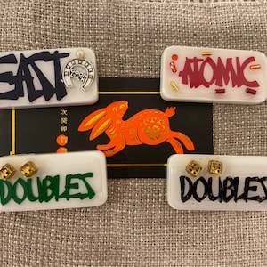 May include: Rectangular white resin pieces with text and embellishments. One says "EAT" with a horseshoe charm. Another says "ATOMIC". Two say "DOUBLES" with dice. A red and gold rabbit graphic is in the background.