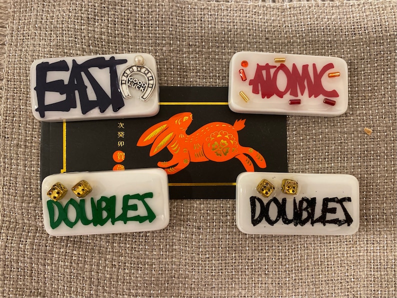 May include: Rectangular white resin pieces with text and embellishments. One says "EAT" with a horseshoe charm. Another says "ATOMIC". Two say "DOUBLES" with dice. A red and gold rabbit graphic is in the background.