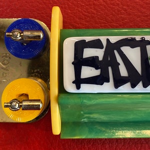 May include: A vintage Royal Games board game component with colorful pieces. The game includes a metal piece with red, blue, green, and yellow circular pieces. A white rectangular piece with the word "EAT" in black is also present.