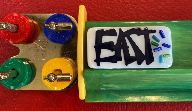 May include: A vintage Royal Games board game component with colorful pieces. The game includes a metal piece with red, blue, green, and yellow circular pieces. A white rectangular piece with the word "EAT" in black is also present.