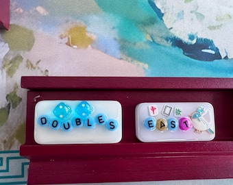 May include: Two white rectangular tiles resting on a dark red wooden rack. The left tile features two blue dice and blue letter beads spelling DOUBLES. The right tile displays small mahjong-style symbols and beads spelling EAST with a bird charm.