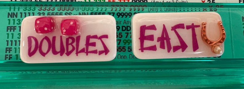 May include: Two rectangular trays with the words "DOUBLES" and "EAST" in magenta lettering. One tray has two red dice with white dots. The other tray has a gold horseshoe charm with a pearl.