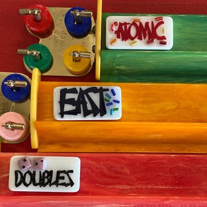 May include: A close-up of a board game with colorful components. The game features tracks in red, orange, and green, with white tiles displaying the words "ATOMIC", "EAT", and "DOUBLES". Dice and colorful game pieces are also visible.