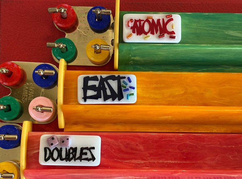 May include: A close-up of a board game with colorful components. The game features tracks in red, orange, and green, with white tiles displaying the words "ATOMIC", "EAT", and "DOUBLES". Dice and colorful game pieces are also visible.