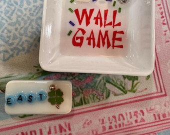 Puede incluir: Un plato cuadrado blanco con las palabras "WALL GAME" en rojo. El plato contiene una ficha de Mahjong, un charm plateado y una pequeña pieza rectangular con la palabra "EAST" y un trébol verde. El fondo es un estampado floral.