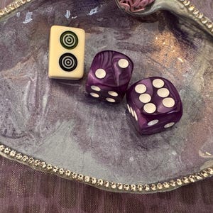 May include: A decorative purple tray with a silver beaded edge holds a cream-coloured tile with black and green concentric circles and two purple dice with white dots. The tray has a marbled design.