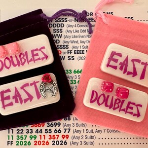 May include: Two sets of game pieces with the words "DOUBLES" and "EAST" in pink on white rectangles. One set is in a purple velvet bag, the other in a pink bag. Each set includes pink dice and a horseshoe charm.