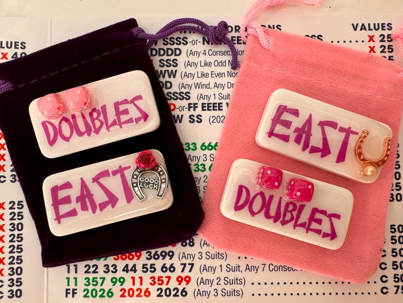 May include: Two sets of game pieces with the words "DOUBLES" and "EAST" in pink on white rectangles. One set is in a purple velvet bag, the other in a pink bag. Each set includes pink dice and a horseshoe charm.