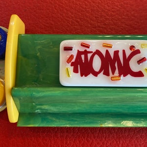 May include: A retro-style Atomic pinball machine component. The white rectangular piece has the word "ATOMIC" in red, with colorful accents. The component is set against a green and yellow background.