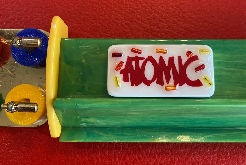 May include: A retro-style Atomic pinball machine component. The white rectangular piece has the word "ATOMIC" in red, with colorful accents. The component is set against a green and yellow background.