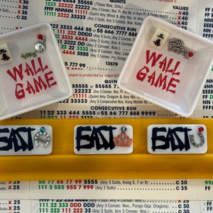 May include: Two small white square dishes with the words "WALL GAME" in red, and decorative charms. Below are three rectangular tiles with the word "EAST" and various charms. The background is a yellow surface with printed text.