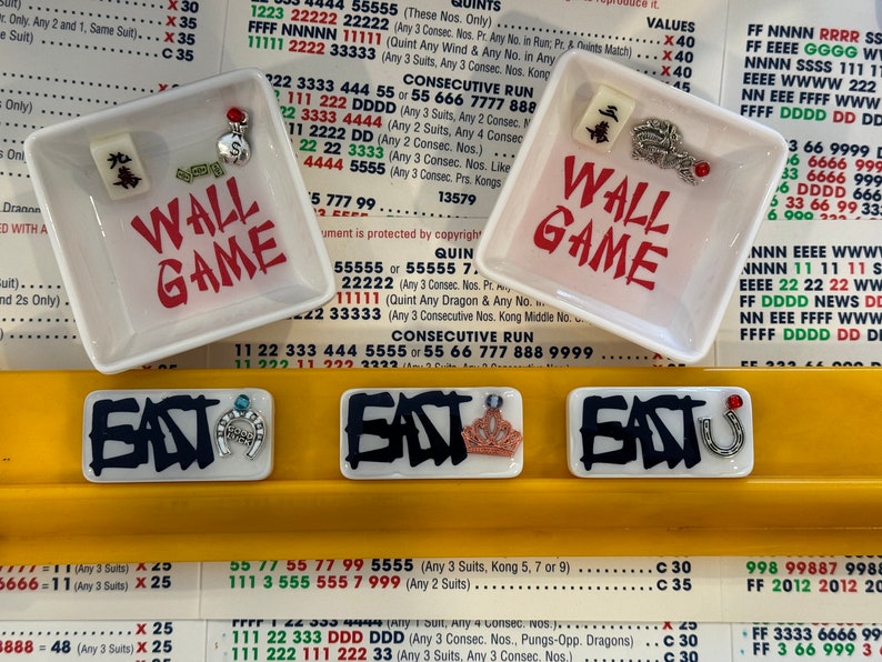 May include: Two small white square dishes with the words "WALL GAME" in red, and decorative charms. Below are three rectangular tiles with the word "EAST" and various charms. The background is a yellow surface with printed text.