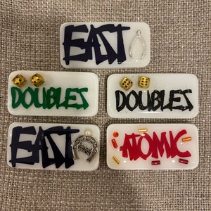 May include: Five rectangular, white resin pieces with various designs. The pieces feature the words "EAT", "DOUBLES", and "ATOMIC" in different colors and styles, along with small charms and dice.