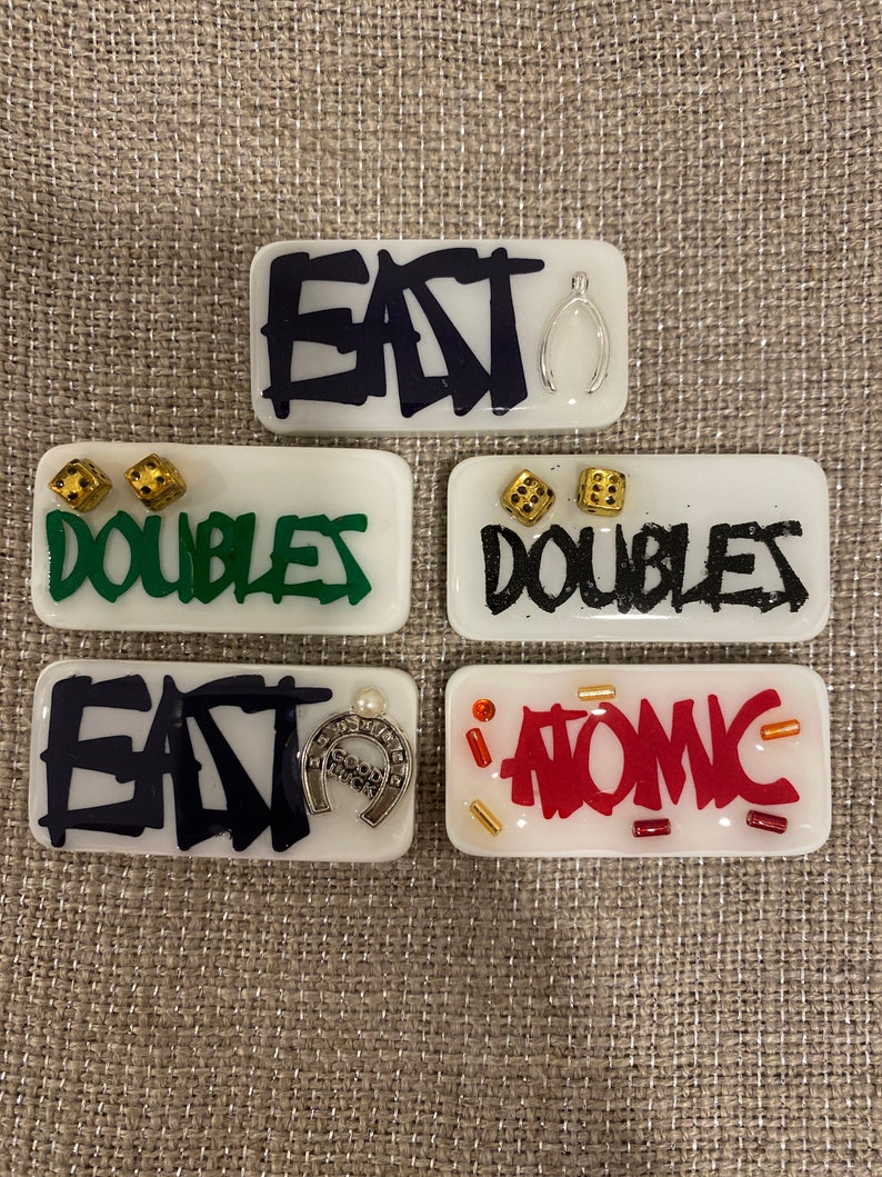 May include: Five rectangular, white resin pieces with various designs. The pieces feature the words "EAT", "DOUBLES", and "ATOMIC" in different colors and styles, along with small charms and dice.