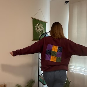 Puede incluir: Una sudadera burdeos con un diseño de patchwork en la espalda, con cuadrados de telas moradas, doradas, verdes y azules. La sudadera tiene mangas largas y cuello redondo. La persona lleva vaqueros azules.
