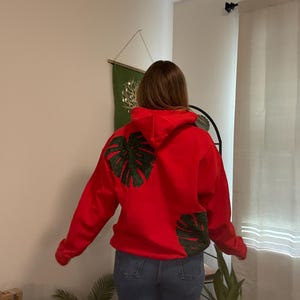 May include: A red hoodie with a green monstera leaf design on the back and sleeve. The hoodie is paired with blue jeans. The person is standing in a room with a neutral-colored wall and a decorative hanging.
